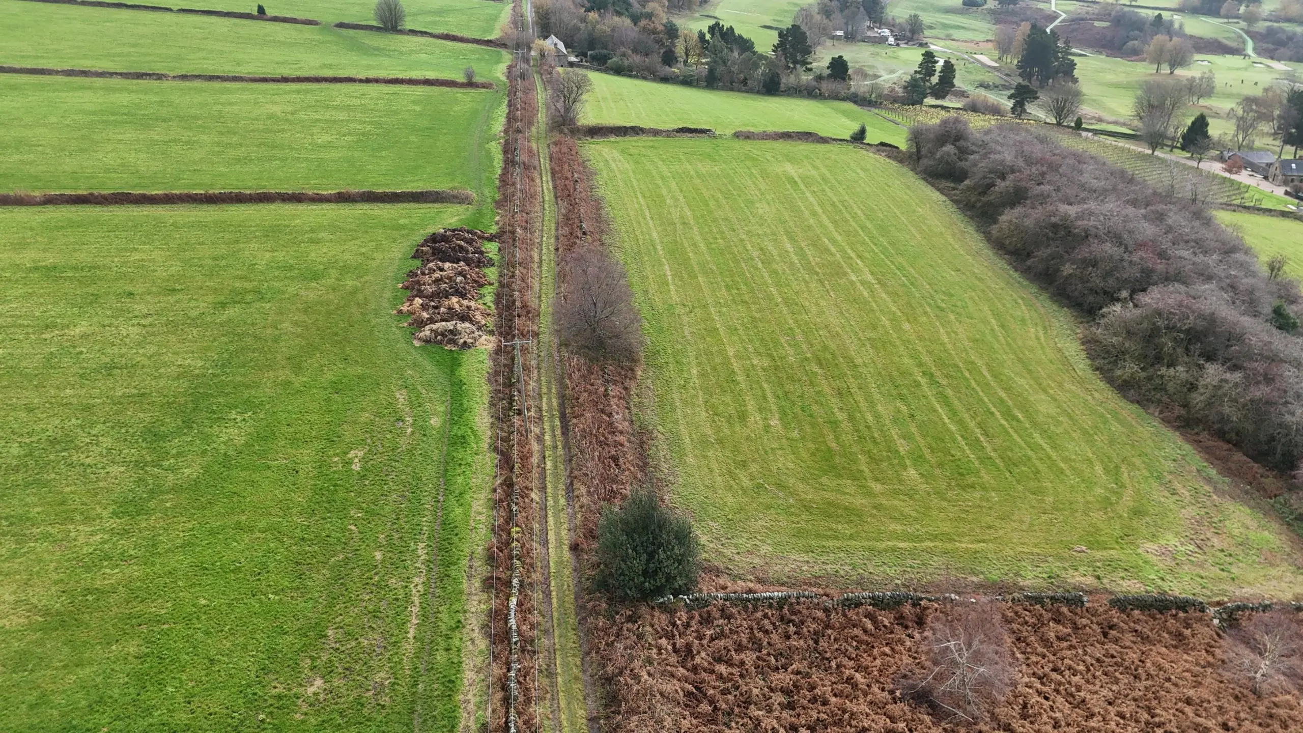 Aerial view of rural farmland with green fields divided by hedgerows and tree lines, ploughed brown field in foreground, scattered buildings and woodland visible in distance